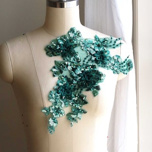Pode incluir: Aplique floral verde-azulado com lantejoulas e contas sobre um fundo de tecido branco.