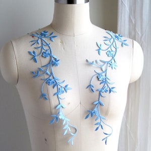 以下が含まれることがあります： 白いマネキンに飾られた、繊細なツルと小さな花が付いた薄い青色の花柄レースのアップリケ2つ