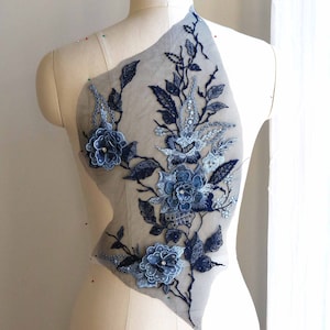 May include: A blue floral lace applique with intricate detailing. The applique features embroidered flowers, leaves, and vines in shades of blue, accented with sequins. The design is mounted on a sheer, gray fabric backing, perfect for embellishing garments.