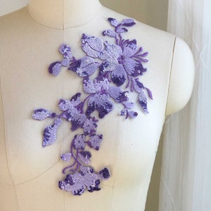 May include: A purple floral applique with sequins and beads. The applique is on a white fabric background.