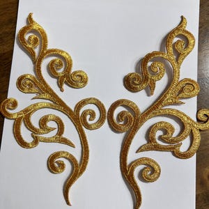 May include: Two gold embroidered appliques with a swirling, floral design. The appliques are symmetrical, with a central stem and curling tendrils. The embroidery has a textured appearance, and the colour is a bright, metallic gold.