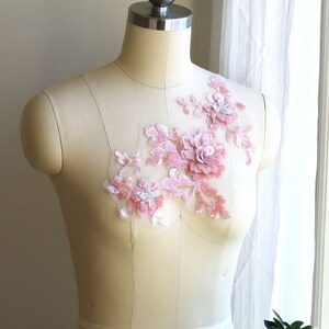 May include: A pink floral applique with sequins and beads on a white mesh fabric. The applique is displayed on a white dress form.