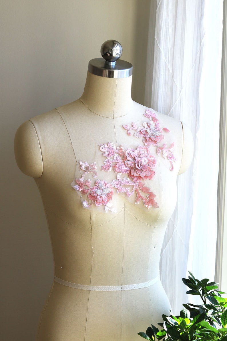 May include: A pink floral applique with sequins and beads on a white mesh fabric. The applique is displayed on a white dress form.
