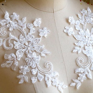 May include: Two white floral lace appliques with intricate detailing. Each applique features a design of flowers, leaves, and swirling patterns, embellished with sequins. These appliques are ideal for bridal wear or other craft projects.