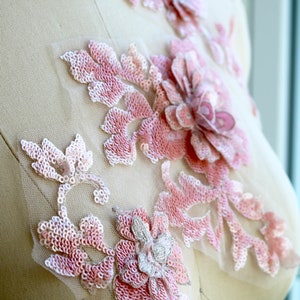 May include: Pink floral applique with sequins and beads on a sheer fabric. The applique features a large flower with smaller flowers and leaves surrounding it.