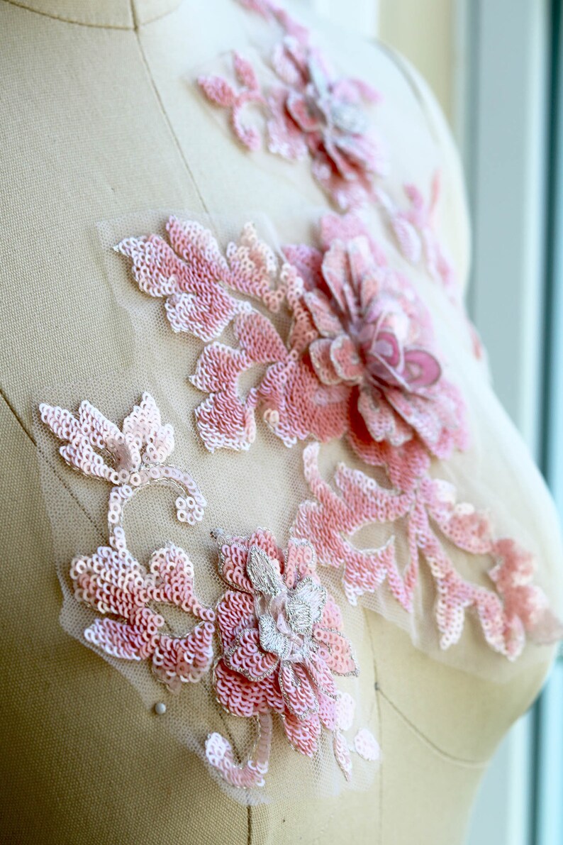 May include: Pink floral applique with sequins and beads on a sheer fabric. The applique features a large flower with smaller flowers and leaves surrounding it.