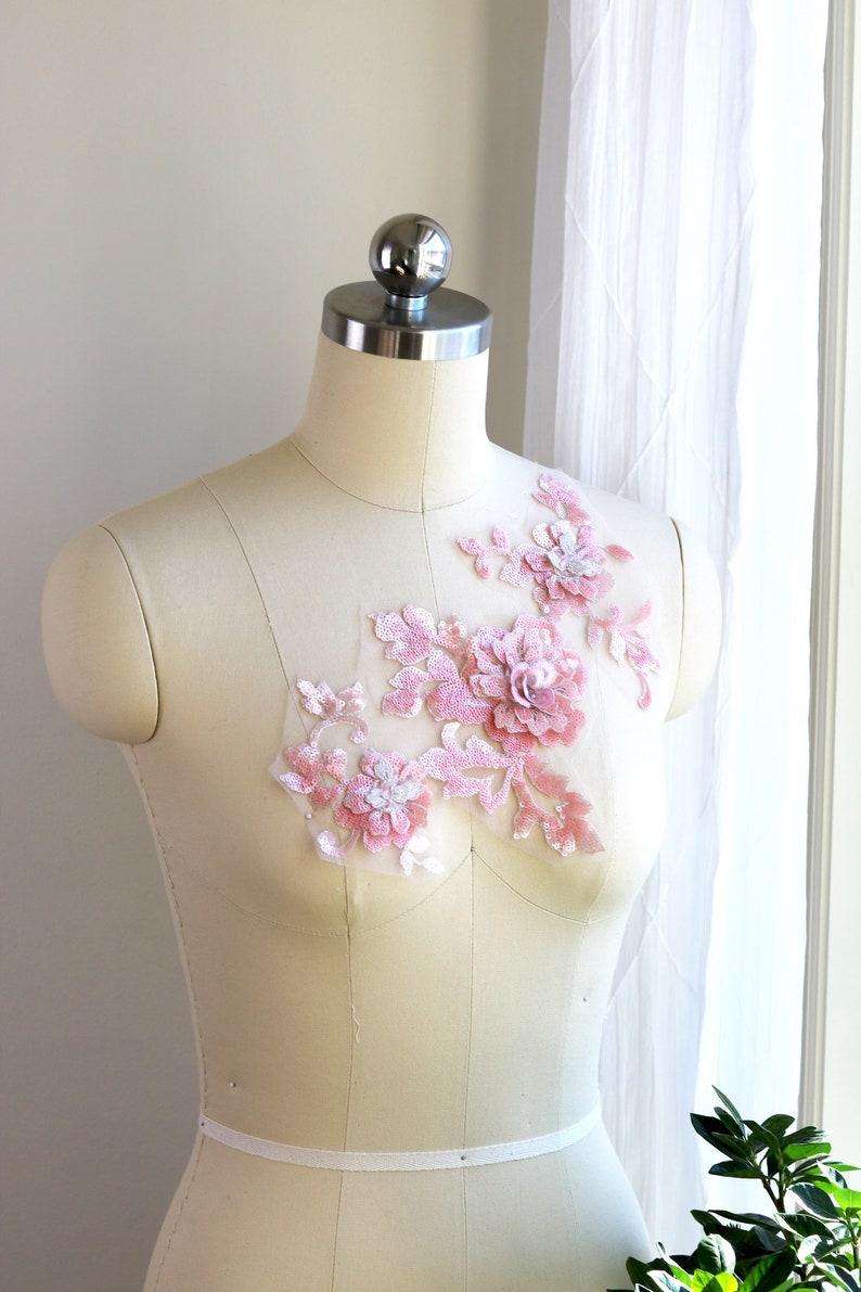 May include: A pink floral applique with sequins and pearls on a white dress form.