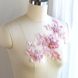 May include: A pink floral applique with sequins and beads on a white fabric background. The applique is perfect for adding a touch of elegance to any garment.