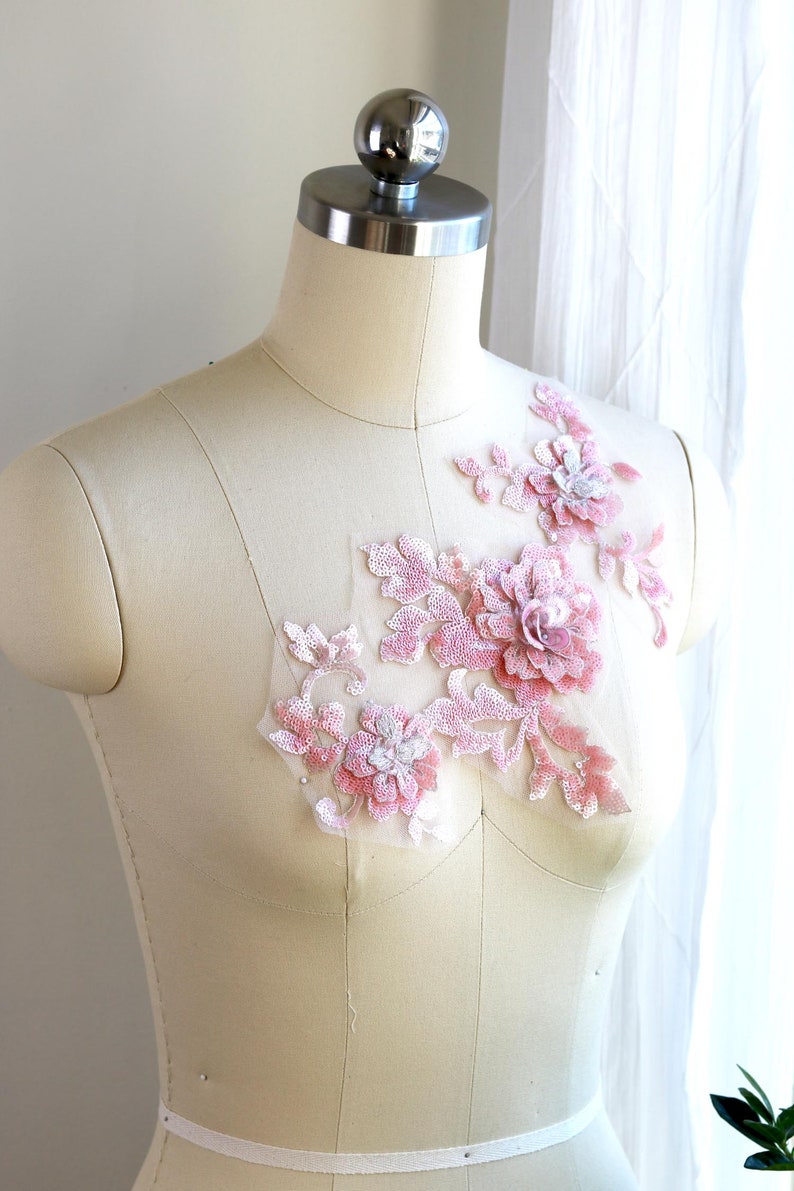 May include: A pink floral applique with sequins and beads on a white fabric background. The applique is perfect for adding a touch of elegance to any garment.