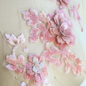 May include: Pink floral applique with sequins and beads. The applique is made of delicate fabric and features a floral design with a large central flower and smaller surrounding flowers. The applique is perfect for adding a touch of elegance to any garment or project.