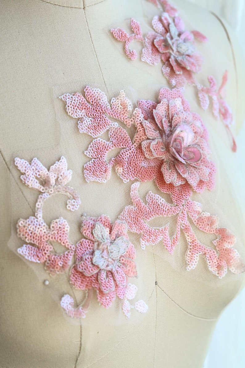 May include: Pink floral applique with sequins and beads. The applique is made of delicate fabric and features a floral design with a large central flower and smaller surrounding flowers. The applique is perfect for adding a touch of elegance to any garment or project.