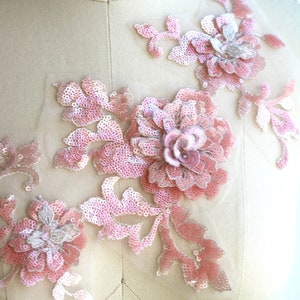 May include: Pink floral applique with sequins and beads. The applique features a large flower with smaller flowers and leaves. The applique is on a white background.