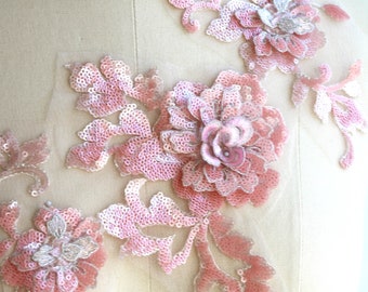 May include: Pink floral applique with sequins and beads. The applique features a large flower with smaller flowers and leaves. The applique is on a white background.