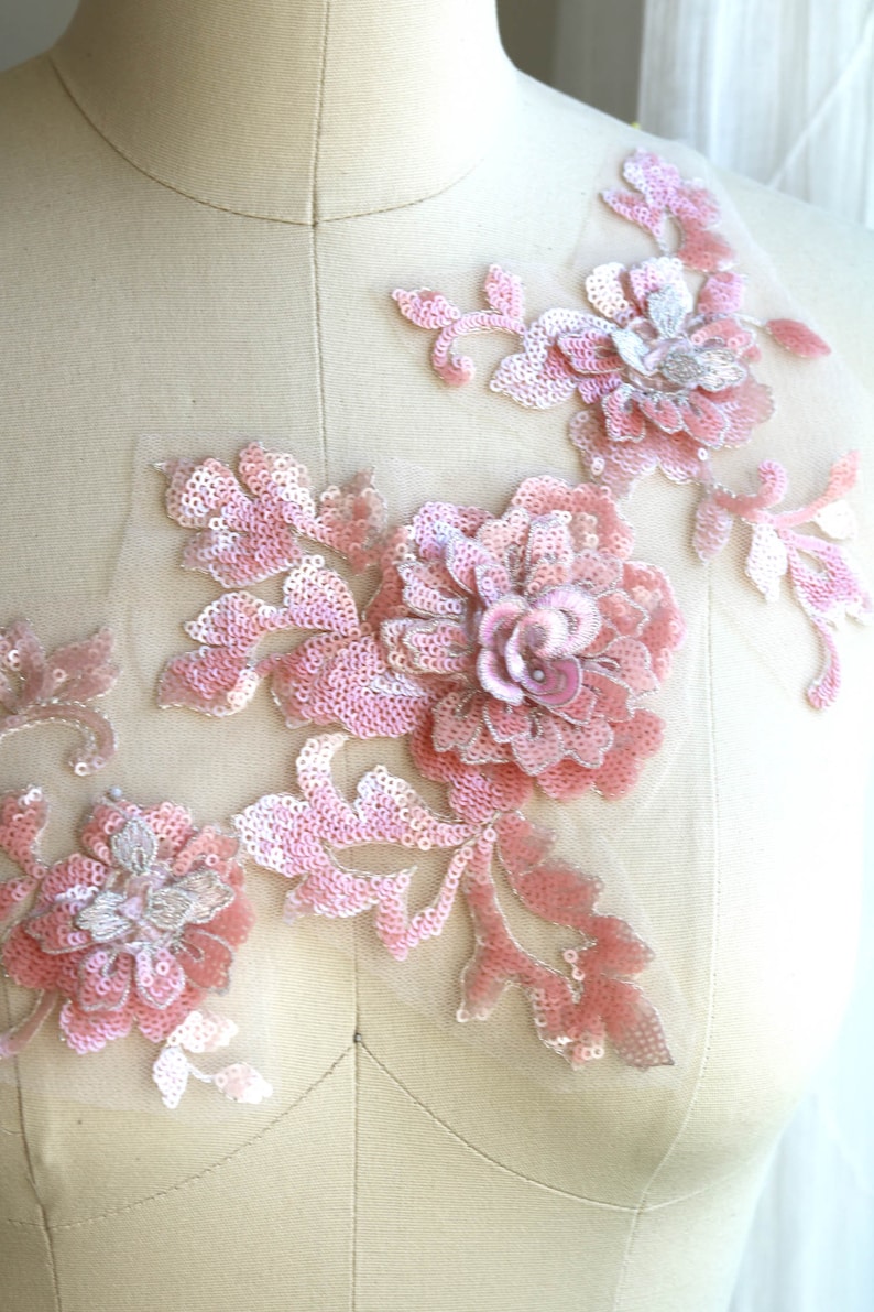 May include: Pink floral applique with sequins and beads. The applique features a large flower with smaller flowers and leaves. The applique is on a white background.