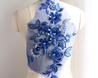 Puede incluir: Aplique floral azul y plateado con lentejuelas y cuentas sobre una tela transparente. El aplique tiene forma de flor y un diseño delicado.