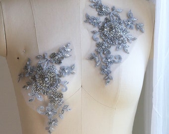 May include: Two silver beaded floral appliques on a white dress form. The appliques are made of silver beads and sequins and have a delicate floral design.