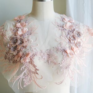 Könnte beinhalten: Eine zarte rosa florale Spitzenapplikation mit Perlenbesatz. Die Spitze befindet sich auf einem durchsichtigen Netzstoff und zeigt 3D-Blumen.