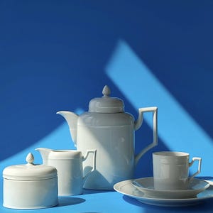 May include: A white porcelain tea set against a blue background. The set includes a teapot, creamer, sugar bowl, teacup, and saucer. The teapot has a lid with a finial, and the teacup has a handle. The set is designed with clean lines and a minimalist aesthetic.