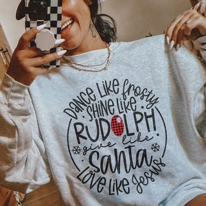 May include: A light gray sweatshirt with the text "Dance Like Frosty, Shine Like Rudolph, Give Like Santa, Love Like Jesus" in a black script. The word "Rudolph" has a red plaid design. The person is wearing a beige cap.