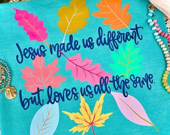 May include: A teal t-shirt featuring the dark blue text Jesus made us different but loves us all the same, surrounded by colorful graphic leaves in shades of pink, orange, yellow, and light blue, with a beaded bracelet and a light pink bow.