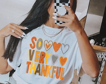 May include: White t-shirt with the words "SO VERY THANKFUL" in orange, yellow, and green, with heart graphics. The shirt is knotted at the waist. The person is wearing checkered shorts.