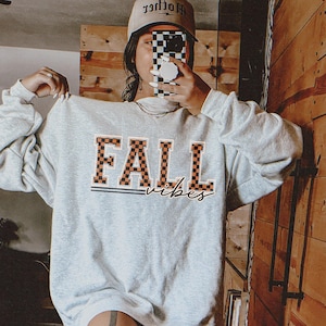 May include: A gray sweatshirt with the text "FALL vibes" in orange and black checkered print. The sweatshirt is being worn by a person.