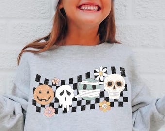 May include: A light grey crewneck sweatshirt featuring a black and white checkered band across the chest. The design includes a pumpkin, a ghost, a mummy, and a skull, accented with small flowers. The sweatshirt has long sleeves and ribbed cuffs.