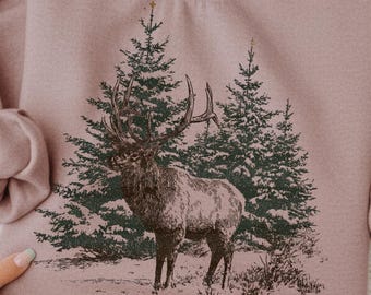 May include: A mauve sweatshirt with a detailed illustration of an elk standing before two evergreen trees. The elk has large antlers. The sweatshirt has long sleeves and a crew neck.