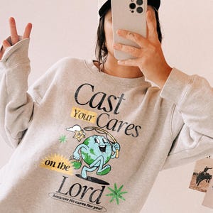 Puede incluir: Una sudadera gris claro con el texto "Cast Your Cares on the Lord" y un gr&aacute;fico de la tierra de dibujos animados. La sudadera tiene mangas largas y cuello redondo. La persona de la imagen lleva una gorra de b&eacute;isbol y sostiene un tel&eacute;fono.