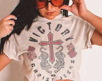 May include: A beige short-sleeved t-shirt featuring a graphic print of a central cross, two tigers, and floral accents. The text "VICTORY IN CHRIST JESUS" is printed in a circular arrangement around the central design in a distressed black font.