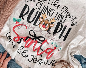 May include: A light gray t-shirt featuring black and red text that reads Dance Like Frosty, Shine Like Rudolph, Give Like Santa, Love Like Jesus. The design includes a cartoon reindeer, a black bow, holly, and colorful Christmas light bulbs.
