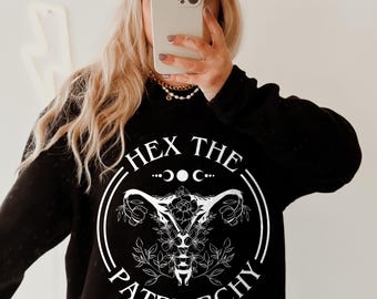 May include: A black long-sleeve sweatshirt featuring a white graphic of a floral uterus design enclosed in a circle with the text "HEX THE PATRIARCHY" and moon phases above it. The garment is paired with a bright orange baseball cap.
