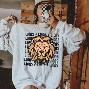 May include: A white sweatshirt with a graphic print of a lion's head. The print is repeated multiple times and says "Lions" in a bold, black font.