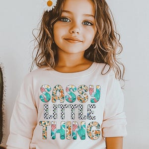 Puede incluir: Una ni&ntilde;a peque&ntilde;a con una camiseta rosa claro de manga larga con las palabras "SASSY LITTLE THING" en un dise&ntilde;o floral y a cuadros. Tiene flores de margarita en el pelo. Se ve un cactus.