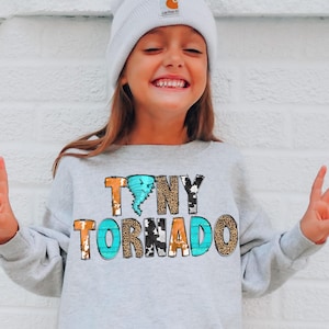 May include: A light gray long-sleeve sweatshirt featuring the text "TINY TORNADO" in a multi-patterned design. The letters incorporate turquoise, leopard print, and cow print textures, with a small turquoise tornado graphic above the word tiny.