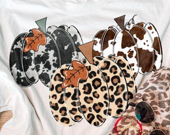 May include: A white short-sleeved t-shirt featuring a graphic of three pumpkins with black and white cow print, brown and white cow print, and leopard print patterns, each topped with a small brown leaf and a stem.