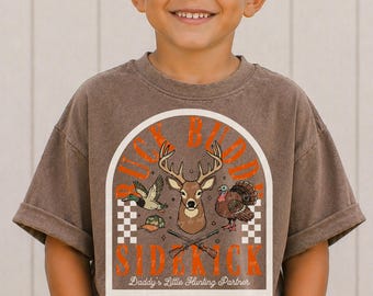 May include: A brown short-sleeve t-shirt featuring a graphic print of a deer head, a duck, a turkey, and two crossed rifles. The text reads "BUCK BUDDY" in orange and "Daddy's Little Hunting Partner" in white, framed by a white arch.