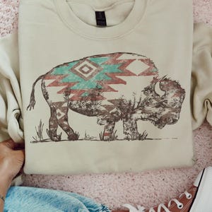 May include: A beige crewneck sweatshirt featuring a large graphic print of a bison. The bison is illustrated with a brown outline and filled with a geometric pattern in shades of teal, red, and white. The sweatshirt has long sleeves and a ribbed collar.