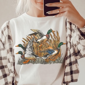 May include: White t-shirt featuring a detailed illustration of four ducks in a wheat field. The ducks have green heads, brown bodies, and blue wing accents. The shirt is worn with a black and white plaid jacket.