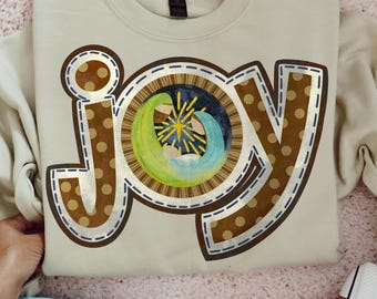 May include: A beige crewneck sweatshirt featuring the word JOY in large brown letters with tan polka dots. The letter O is replaced by a circular graphic depicting a nativity scene with a bright yellow star, green and blue figures, and a dark background.