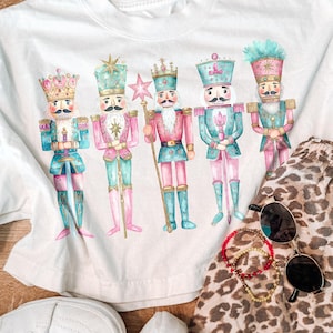 May include: A white t-shirt featuring a graphic print of five nutcracker figures in pastel pink and teal uniforms with gold accents. The nutcrackers are arranged in a row, each wearing a decorative hat and holding a staff or sword.