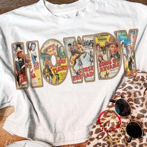 May include: A white short-sleeved t-shirt featuring the word HOWDY in large letters, each filled with vintage Western movie poster graphics. The shirt is paired with leopard print shorts, a pair of black sunglasses, and two beaded bracelets.