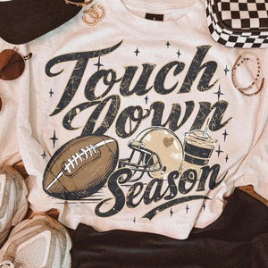 May include: A white t-shirt featuring the text Touch Down Season in dark blue distressed lettering. The graphic includes a brown football, a tan football helmet, and a drink cup, surrounded by small star icons. The shirt is surrounded by accessories.