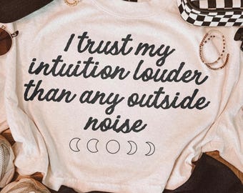 May include: A cream-colored crewneck sweatshirt featuring the black text "I trust my intuition louder than any outside noise" and a series of five moon phase icons. The garment is surrounded by a black and white cap, sunglasses, and sneakers.