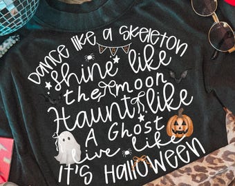 May include: A black t-shirt featuring white text that reads: Dance like a skeleton, shine like the moon, haunt like a ghost, live like it's Halloween. The design includes small illustrations of a spider, a bat, a pumpkin, and a ghost.