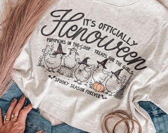 May include: A light gray t-shirt featuring black text that reads It's officially Henoween, Pumpkins in the coop, Treats for the girls, and Spooky season forever. The graphic includes five chickens wearing witch hats and a small orange pumpkin.