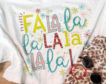 May include: A white short-sleeved t-shirt featuring the colourful text "FA LA LA LA LA LA LA" in various fonts and shades of yellow, pink, red, and teal, accented with small painted snowflakes and dots, paired with leopard print shorts and sunglasses.