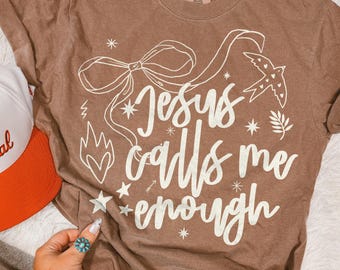 May include: A brown short-sleeve t-shirt featuring white screen-printed text that reads Jesus calls me enough, accompanied by graphic illustrations of a large bow, a bird, a flame, and several small stars on the front of the garment.