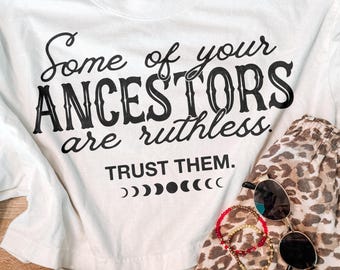 May include: A white t-shirt with black text reading Some of your ANCESTORS are ruthless. TRUST THEM. with a moon phase graphic, paired with leopard print shorts, white sneakers, a pair of black sunglasses, and two beaded bracelets.