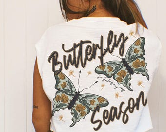 May include: A white short-sleeved crop top featuring a graphic print of two butterflies with muted green and gold floral patterns. The text reads Butterfly Season and I'm finding my wings in a dark brown script font, accompanied by small dandelion illustrations.